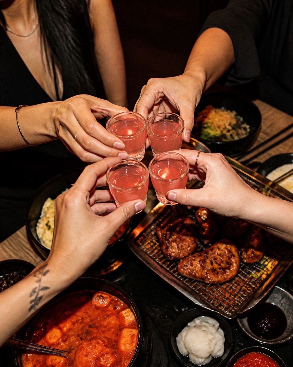 Friends toasting coral-pink KTOWN soju shots over a Korean dinner